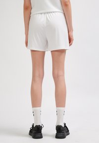 Białe sportowe spodenki z elastycznym paskiem w pasie, gładką strukturą i luźnym krojem. Noszone z czarnymi sportowymi butami i białymi skarpetkami w wzory.