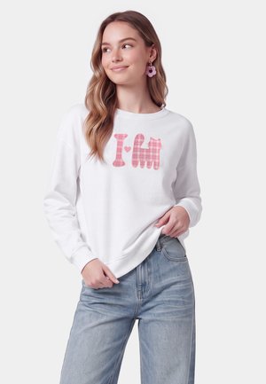 Ung kvinde med langt hår iført hvid sweatshirt med pink kattegrafik og blå jeans, smilende og kiggede til siden.