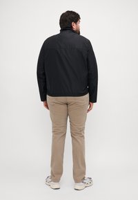 Giacca leggera nera con colletto alto e maniche lunghe, caratterizzata da una silhouette dritta e un design minimalista. Abbinata a pantaloni beige.