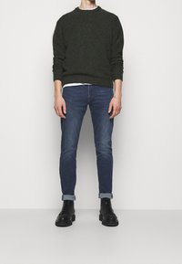 Man i mörkgrön stickad tröja, blå uppvikta jeans och svarta snörade stövlar som står mot en enfärgad vit bakgrund.