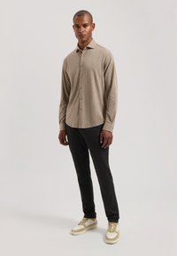 Cămașă bej cu mânecă lungă și guler cu nasturi, combinată cu pantaloni negri și adidași deschiși. Material neted, design ajustat.