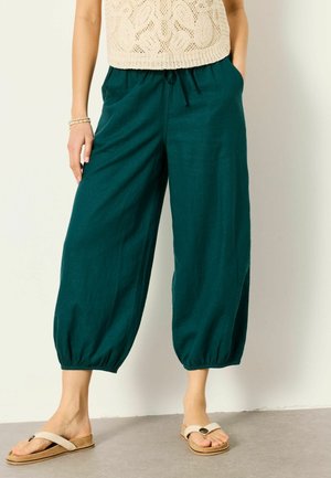 Frau trägt türkisfarbene, weite, verkürzte Hosen mit elastischen Bündchen, ein beige gehäkeltes Oberteil und beigefarbene Sandalen, steht mit den Händen in den Taschen.