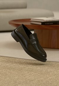 Mocassin en cuir noir flottant au-dessus d'un tapis beige texturé avec une table basse en bois et des livres empilés en arrière-plan.