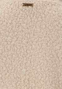 Cremefarbener Stoff mit Boucle-Oberfläche, der kleine, lockige Fasern aufweist. Ein metallisches Etikett ist oben sichtbar.