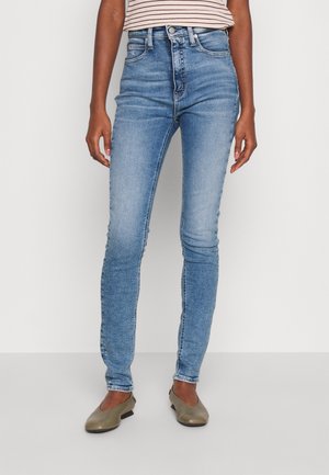 Personne portant un jean skinny bleu clair, des ballerines beiges et un top rayé à manches courtes, posant devant un fond uni.
