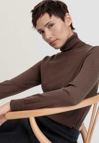 Pull à col roulé marron en matière douce, design ajusté, avec des manches longues. Le mannequin repose un bras sur une chaise en bois clair, texture visible.