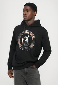 Joven vestido con una sudadera negra con capucha que tiene un logotipo circular multicolor y el texto "Just Cavalli", acompañado de unos pantalones vaqueros gris oscuro, de pie contra un fondo blanco.