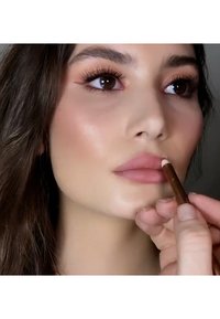 Una matita per labbra viene applicata sulle labbra di un modello; la matita è marrone, e le labbra sono di un rosa delicato con una texture liscia.