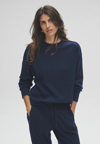 Mujer con cabello largo y castaño que lleva un suéter de canalé azul marino y pantalones a juego, de pie con las manos en los bolsillos sobre un fondo liso.