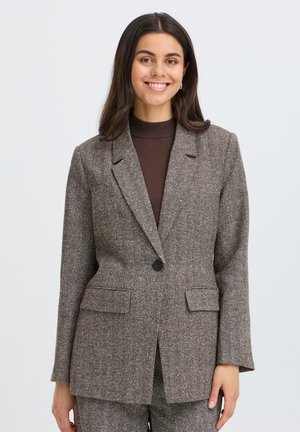Jeune femme souriante, portant un blazer gris à chevrons par-dessus un haut marron foncé, debout devant un fond clair uni.
