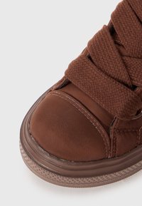 Zapatilla alta marrón con puntera redondeada, parte superior de tela texturizada, cordones gruesos y suela de goma con un dibujo en el desgaste.