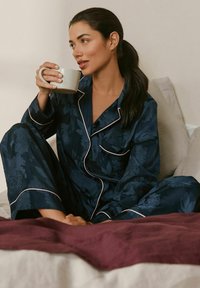Navy floral pajama set with white piping, featuring long sleeves and wide pants. Model holds a white mug while sitting on a bed with maroon bedding.