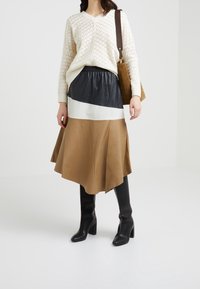 Maglione di maglia crema, gonna in pelle nera e color block in tonalità marrone, stivali neri con tacco al ginocchio, borsa beige, presentati su uno sfondo neutro.