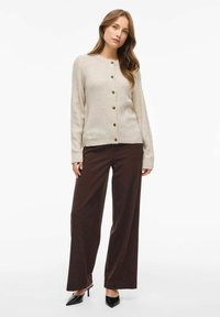 Cárdigan de punto beige con cuello redondo, cierre de botones y mangas largas; combinado con pantalones de pierna ancha marrones y zapatos de tacón negro.