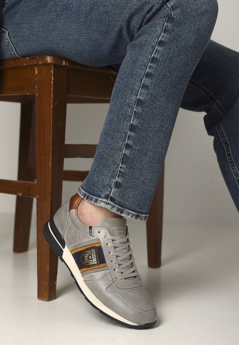 Šedé tenisky s semišovými a koženými akcenty, zdobené zlatým emblémem, šněrováním a strukturovanou černou gumovou podrážkou, nošené s modrými džíny.