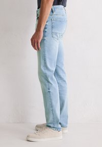 Personne se tenant de côté, portant un jean bleu clair, une chemise noire et des baskets beiges devant un fond blanc uni.