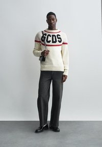 Kremgenser med svarte og røde striper, med fet tekst, paret med mørkegrå vide bukser og svarte sko. Svart veske slengt over skulderen.