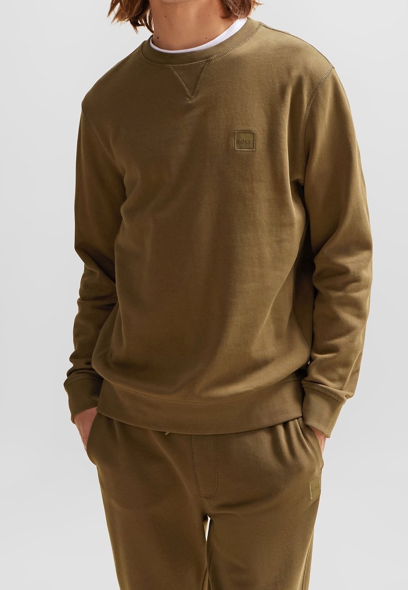 Jeune personne portant un sweat-shirt et un pantalon de survêtement assortis de couleur vert olive, les mains dans les poches, sur un fond uni.