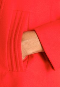 Gros plan sur un manteau en laine rouge montrant une main à l'intérieur d'une poche avec des détails de couture verticale. Le tissu semble doux et texturé.