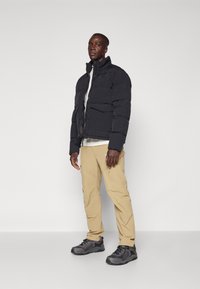 Veste puffer noire avec un col montant, pantalon beige et chaussures de randonnée grises. La veste présente une fermeture éclair et une texture matelassée.