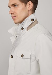 Veste blanche avec un col structuré, des accents de boutons et deux poches poitrine. Présente une texture contrastante au niveau du col et un tissu lisse.