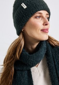 Tmavě zelená pletená čepice a odpovídající šála vyrobená z měkkého, nadýchaného materiálu. Šála má široký design, zatímco čepice obsahuje štítek.