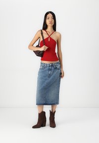 Top sin mangas rojo acanalado con un detalle de ojal, combinado con una falda midi de mezclilla azul. Botines marrones y un pequeño bolso oscuro completan el look.
