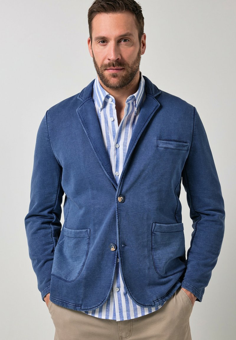 Uomo con capelli castani corti e barba, indossa un blazer blu sopra una camicia a righe blu e bianche, in piedi con le mani in tasca davanti a uno sfondo neutro.