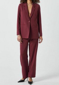 Costume sur mesure bordeaux comprenant un blazer structuré à revers crantés et deux poches avant, associé à un pantalon large assorti et des talons noirs.