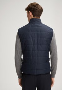 Granatowa pikowana kamizelka z podwyższonym kołnierzem, pikowanym wzorem, bocznymi guzikami i lekką konstrukcją, noszona na szarym topie z długim rękawem.