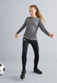 Mergaitė su kuoduku, apsirengusi pilką ilgomis rankovėmis marškinėlį ir juodas tamprias kelnes, žaidžianti su juoda-balta futbolo kamuoliu patalpose.