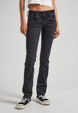 Czarne jeansy typu slim fit noszone z czarno-białymi sneakersami i krótkim białym topem na stojącej osobie.