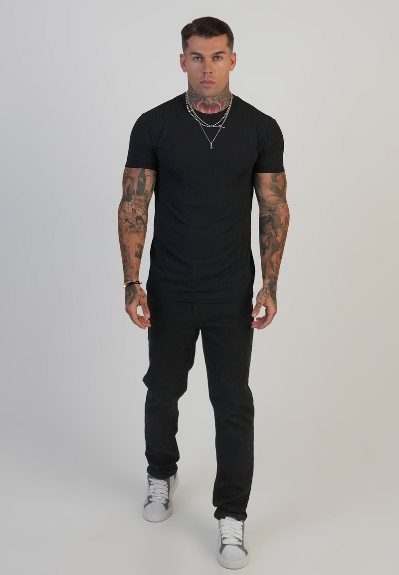 Mann mit Tattoos an Armen und Hals, der ein schwarzes T-Shirt, schwarze Hose, weiße Sneakers und geschichtete silberne Halsketten trägt und nach vorne schaut.