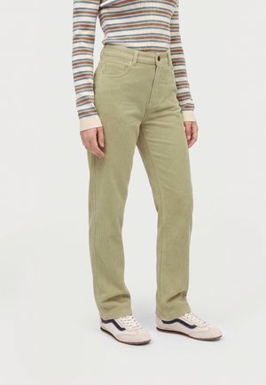 Personne portant un pantalon en velours côtelé vert clair, un haut à manches longues rayé et des baskets blanches avec des détails noirs, debout sur un fond uni.