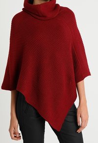 Femme portant un poncho rouge en tricot avec un large col châle, associé à un pantalon noir, debout devant un fond uni.