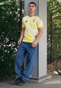 Geel en roze gradient Adidas shirt met drie stripes, gecombineerd met losse blauwe denim jeans en zwarte laarzen. Staande tegen een paal.