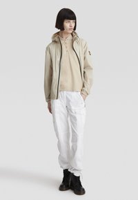 Giacca leggera beige con cappuccio, sopra una polo color cammello, abbinata a pantaloni cargo bianchi e stivali neri con lacci. Texture liscia, vestibilità flessibile.