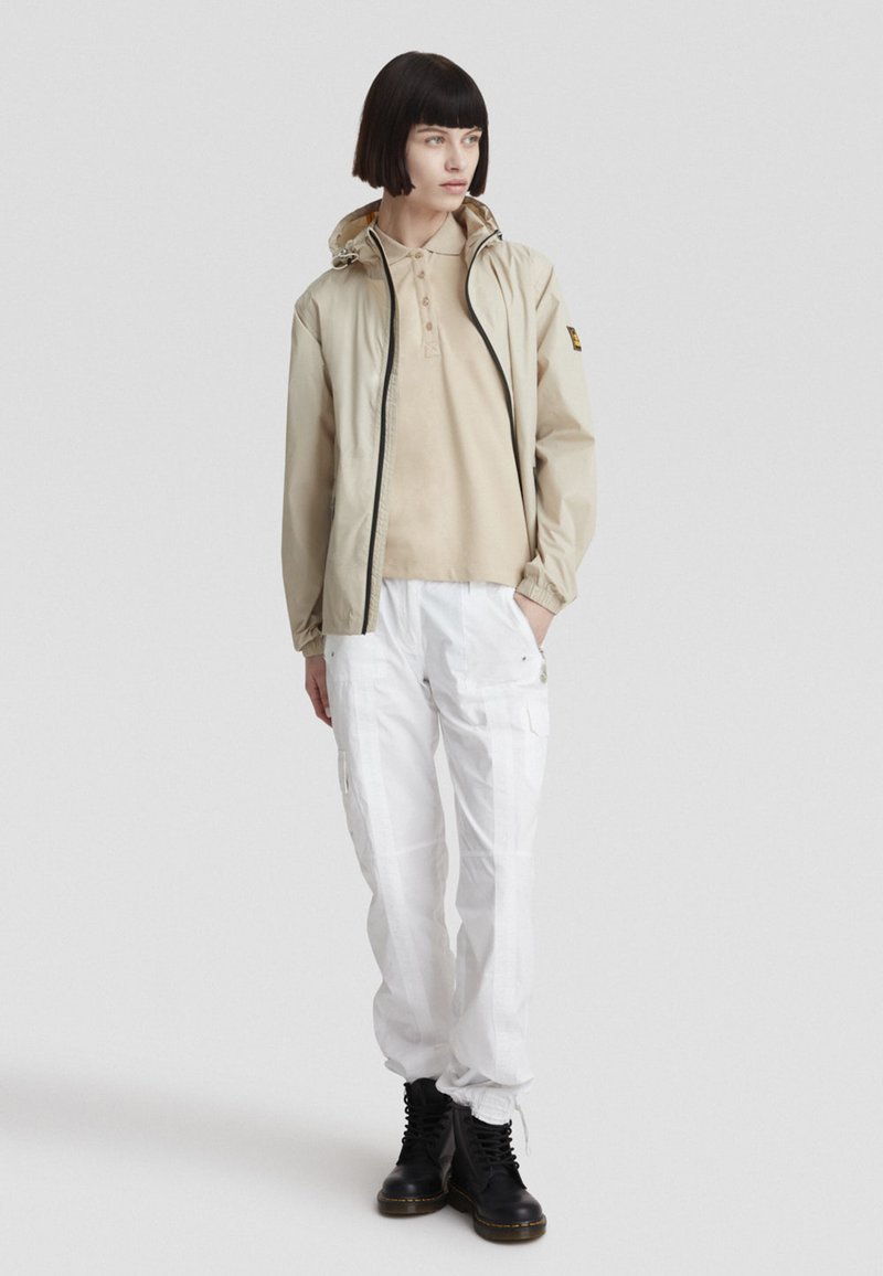 Giacca leggera beige con cappuccio, sopra una polo color cammello, abbinata a pantaloni cargo bianchi e stivali neri con lacci. Texture liscia, vestibilità flessibile.