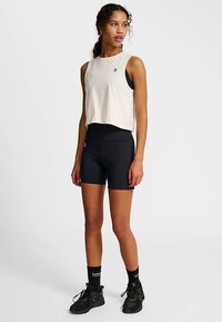 Cropped beige sleeveless top with a small logo, paired with black fitted shorts and black athletic shoes with textured soles.