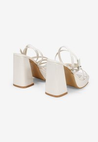 Sandalias blancas de tacón alto con tacones de bloque, que presentan múltiples correas cruzadas y un acabado suave y brillante.