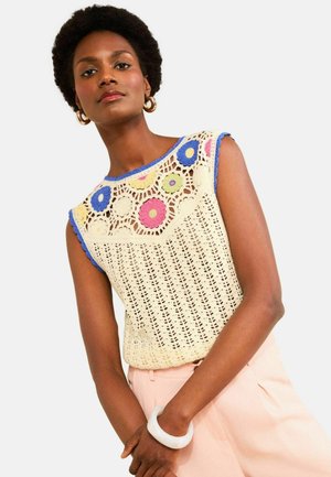 Kvinde iført cremefarvet hæklede ærmeløse top med farverige cirkulære blomster mønstre, blå kant, pink bukser, gyldne hoop øreringe og hvidt armbånd.