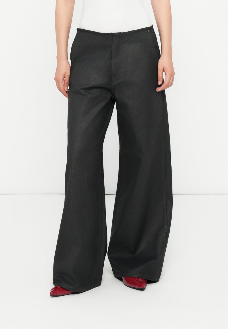 Personne portant un pantalon noir taille haute à jambes larges, associé à des chaussures rouges pointues, debout devant un fond blanc uni.