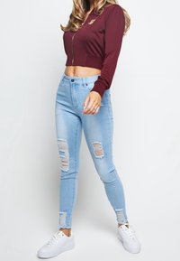 Bordowa krótka kurtka zapinana na zamek z złotymi akcentami, zestawiona z jasnoniebieskimi, zniszczonymi rurkami z dziurami oraz białymi sneakersami.