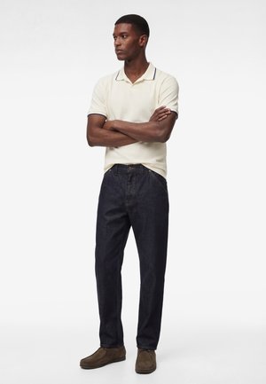 Man staat met gekruiste armen, draagt een witte polo met korte mouwen, donkere jeans en bruine schoenen tegen een effen witte achtergrond.