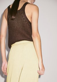 Bruine gehaakte tanktop met een lose fit, gecombineerd met een gele geplooide rok, die een textuurstof en een open rugontwerp toont.