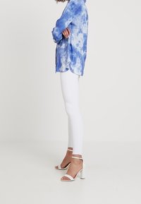 Chemise à teinture nouée bleue en tissu léger, associée à un pantalon skinny blanc et des sandales blanches à talons avec des brides à la cheville.