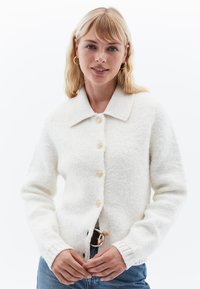 Vit stickad kavaj med krage, fem runda knappar och ribbade ärmslut. Mjuk textur och avslappnad passform. Bärs över blå jeans.