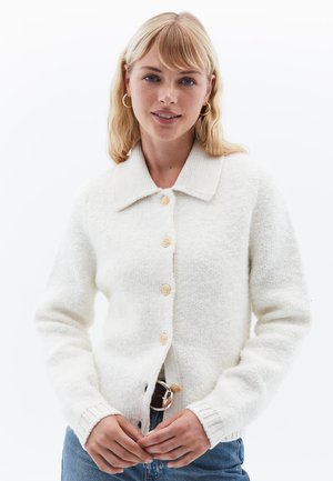Weißer, gestrickter Cardigan mit Kragen, fünf runden Knöpfen und gerippten Bündchen. Weicher Stoff und lockere Passform. Über blauen Jeans getragen.