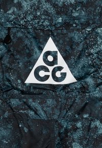 Svart och teal-mönstrad tyg med en triangulär vit logotyp som har bokstäverna "a", "c" och "g" i fet stil. Texturerad yta med sömnadsdetaljer.