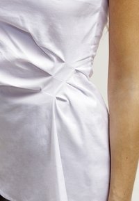 Close-up van een wit shirt met geplooide stofdetails aan de zijkant van de taille, naast een blote arm met zichtbare huidstructuur.
